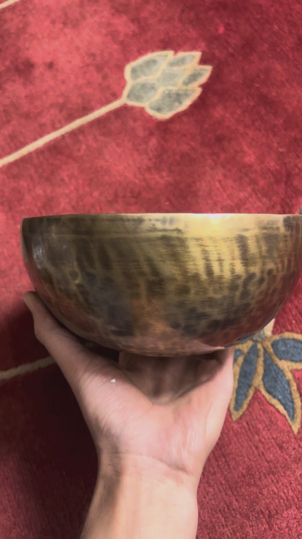 Load video: Side profile view of a small golden bronze Tiger style hand-hammered singing bowl with mottled surface patina, resting on a blue brocade cushion.