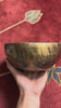 Side profile view of a small golden bronze Tiger style hand-hammered singing bowl with mottled surface patina, resting on a blue brocade cushion.