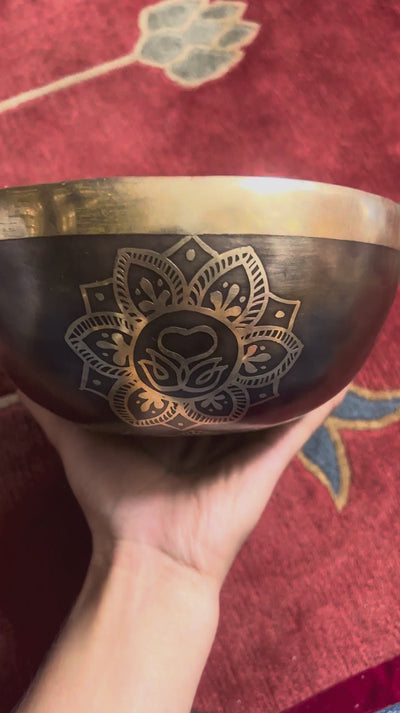 Dark antique bronze Tibetan singing bowl with a golden Swan Symbol of Life engraving on the exterior, displayed on a blue brocade cushion.