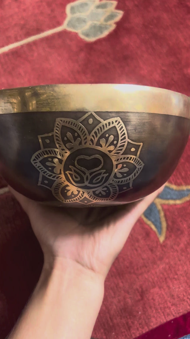 Load video: Dark antique bronze Tibetan singing bowl with a golden Swan Symbol of Life engraving on the exterior, displayed on a blue brocade cushion.
