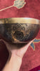 Dark antique bronze Tibetan singing bowl with a golden Swan Symbol of Life engraving on the exterior, displayed on a blue brocade cushion.