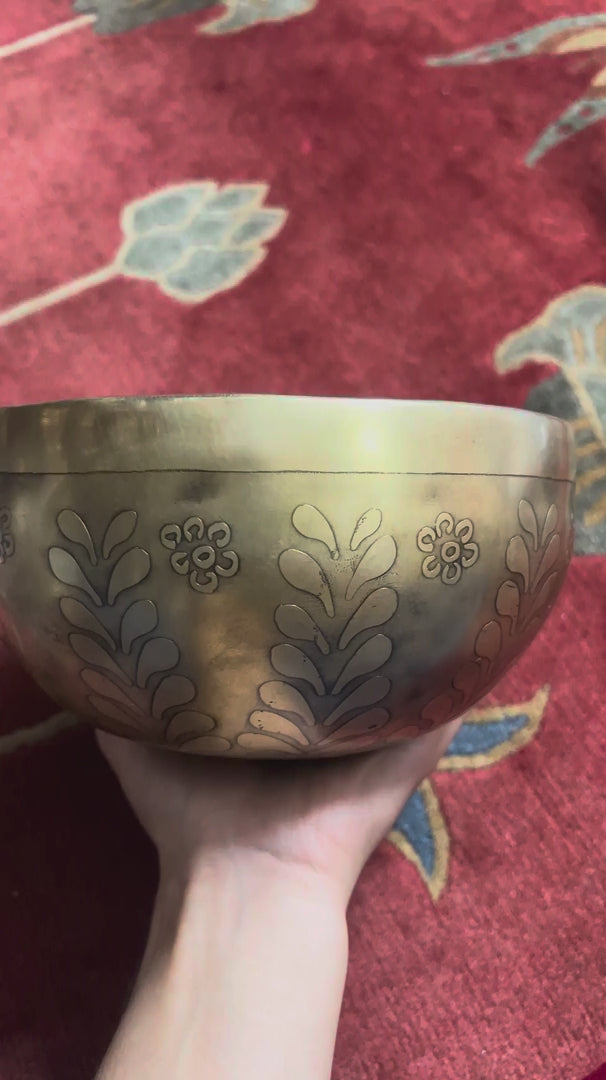 Load video: Antique golden Tree of Life Tibetan singing bowl with engraved leaf and floral patterns displayed on a blue brocade cushion.