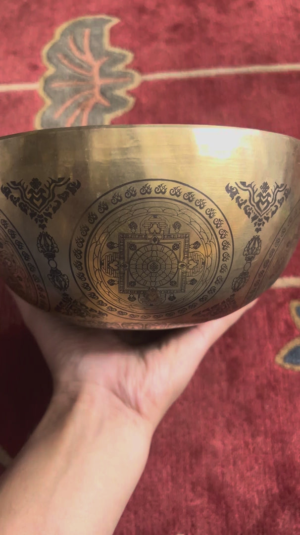 Load video: Engraved Seven Chakra Singing Bowl displayed upright with a wooden mallet and blue cushion, showing the central meditation design.