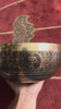 Engraved Tibetan singing bowl featuring a Flower of Life design, photographed upright with a wooden mallet and blue brocade cushion.