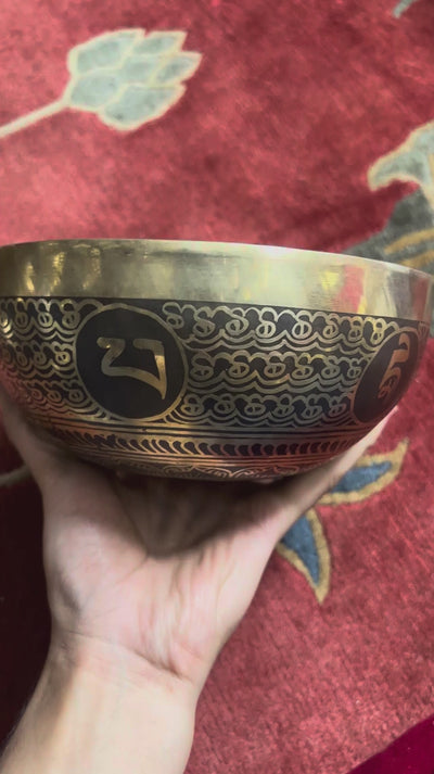 Engraved Endless Knot singing bowl displayed upright on a blue brocade cushion beside a wooden mallet, showing the central symbol and ornate interior borders.