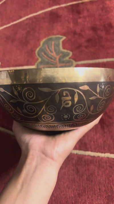 Tree of Life engraved singing bowl displayed upright with a wooden mallet and blue brocade cushion on a woven mat.