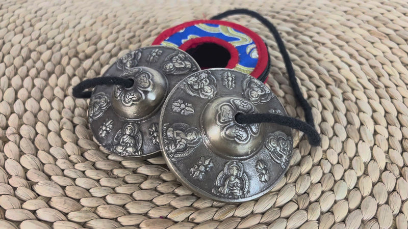 Tibetan Buddha tingsha cymbals featuring detailed Buddha engravings, a black connecting cord, and a colorful brocade carrying case.