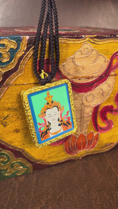 Square gold-framed Vajrasattva pendant with blue background, held in an open hand against a black backdrop.
