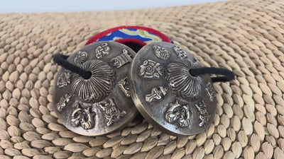 Angled view of a pair of Tibetan tingsha cymbals showing raised Buddhist motifs and decorative central details.