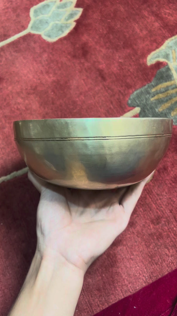 Side profile view of a small warm golden hand-hammered bronze singing bowl with double rim lines, displayed on a blue brocade cushion.