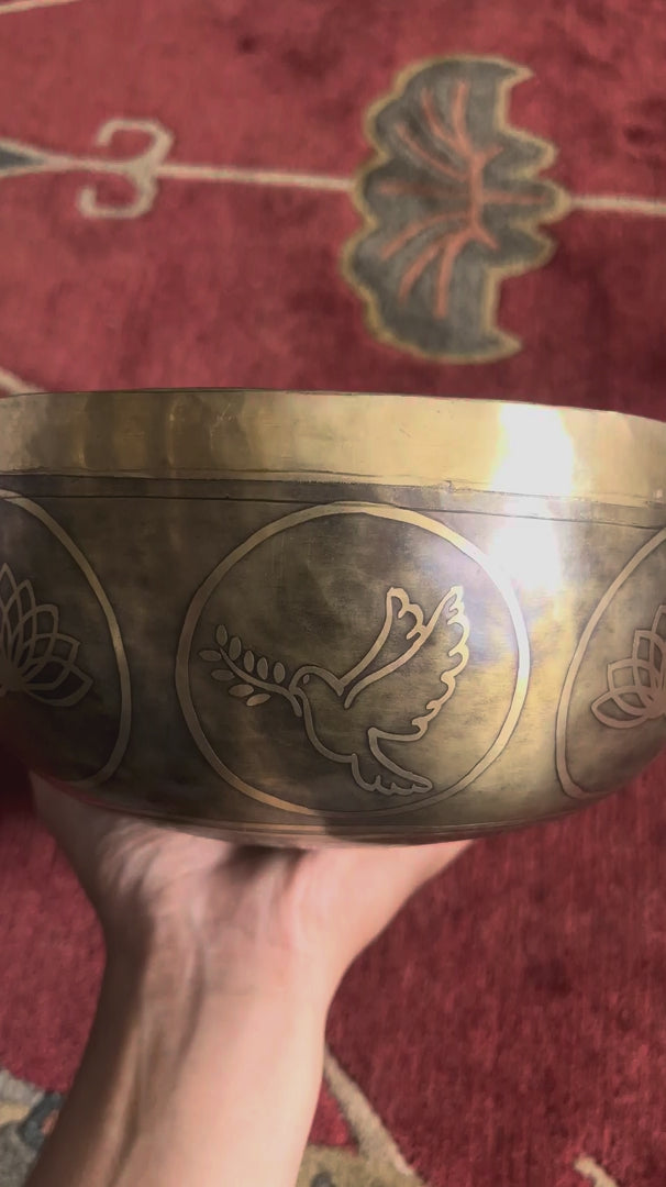 Load video: Golden bronze Dove Tibetan singing bowl featuring engraved dove and lotus flower designs on a blue brocade cushion.