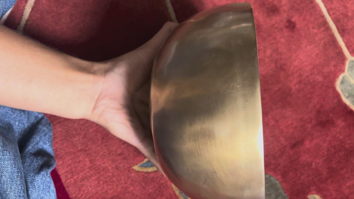 Load video: Side view of a classic plain hand-hammered bronze Tibetan singing bowl resting on a blue brocade cushion.