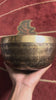 Singing bowl displayed upright with a wooden mallet and blue brocade cushion, showing the central Flower of Life geometric pattern and Tibetan script.