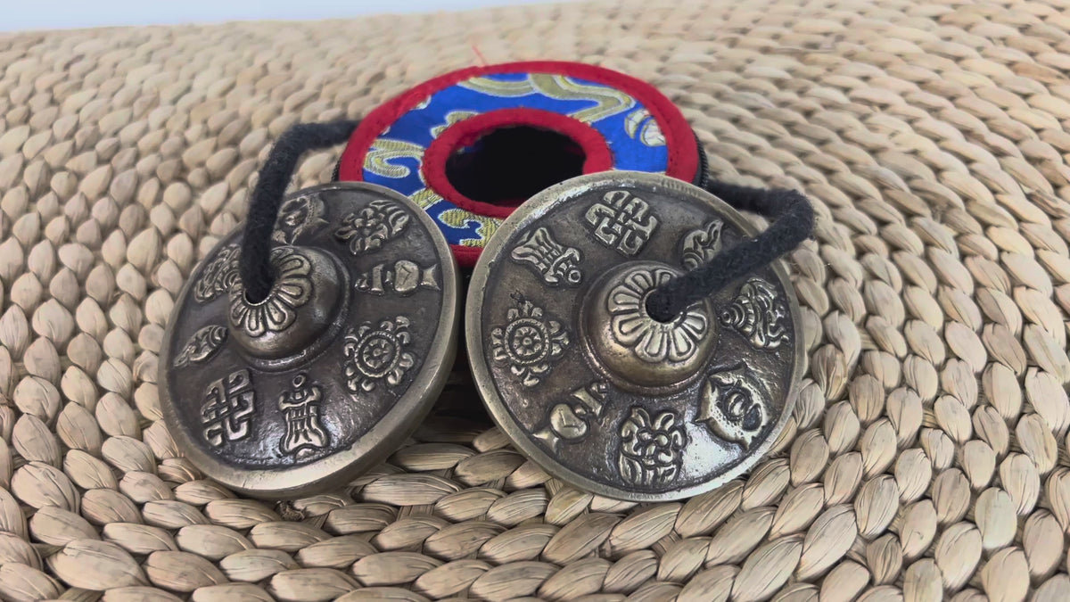 Load video: Pair of Tibetan Buddhist tingsha cymbals with eight auspicious symbols displayed beside a colorful brocade case.