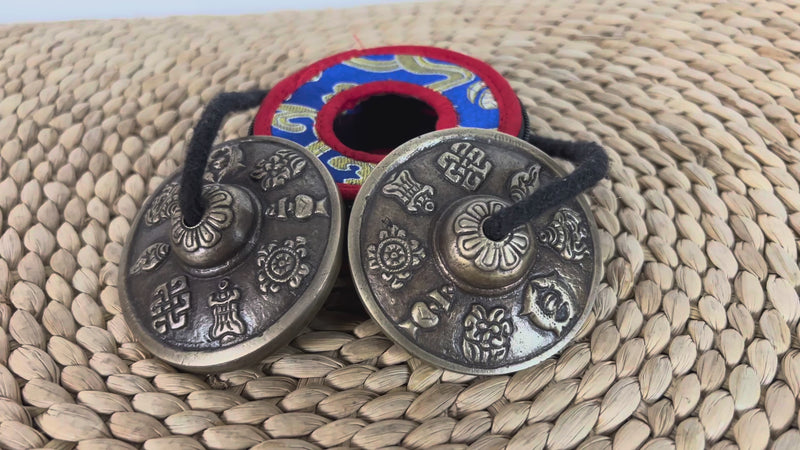 Pair of Tibetan Buddhist tingsha cymbals with eight auspicious symbols displayed beside a colorful brocade case.