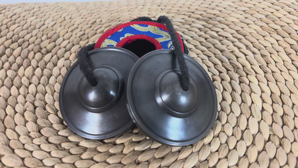 Load video: Traditional black Tibetan ritual cymbals photographed beside a colorful brocade carrying pouch.