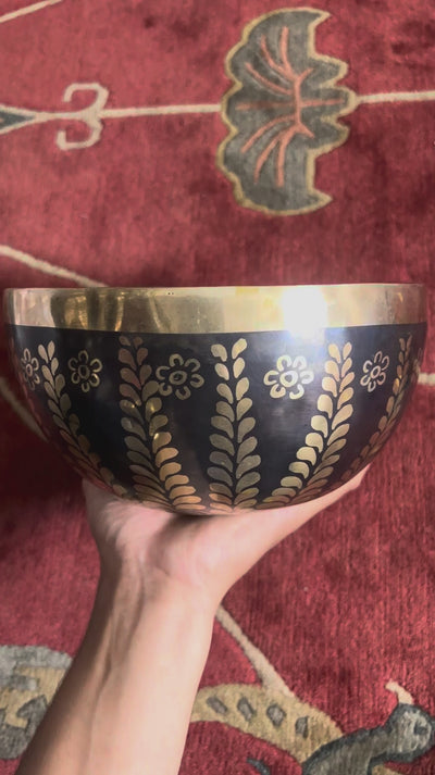 Dark bronze Tibetan singing bowl with golden Tree of Life inspired leaf and floral engravings displayed on a blue brocade cushion.