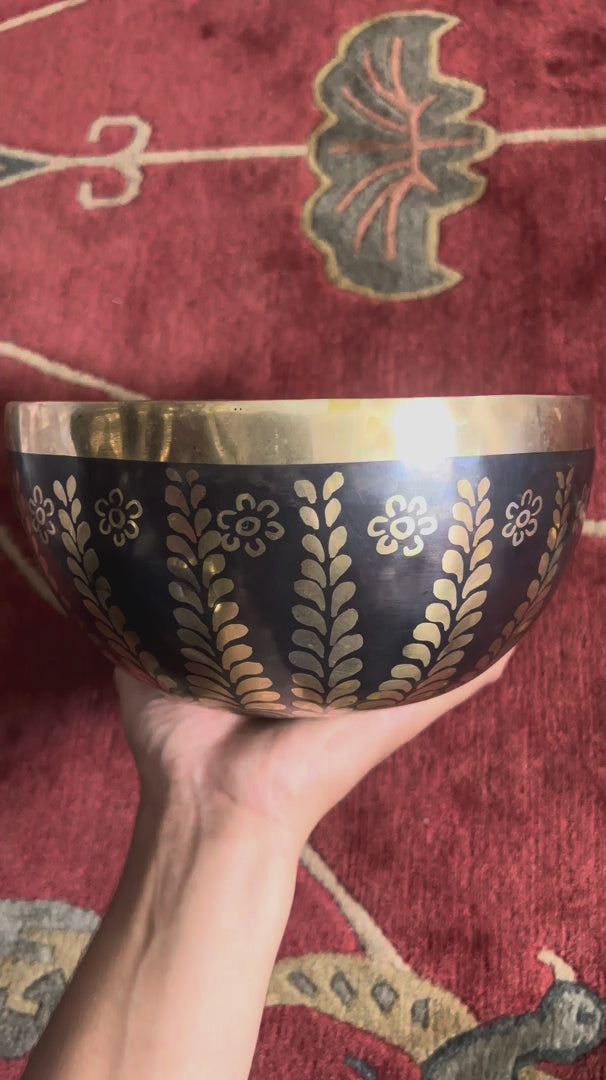 Load video: Dark bronze Tibetan singing bowl with golden Tree of Life inspired leaf and floral engravings displayed on a blue brocade cushion.